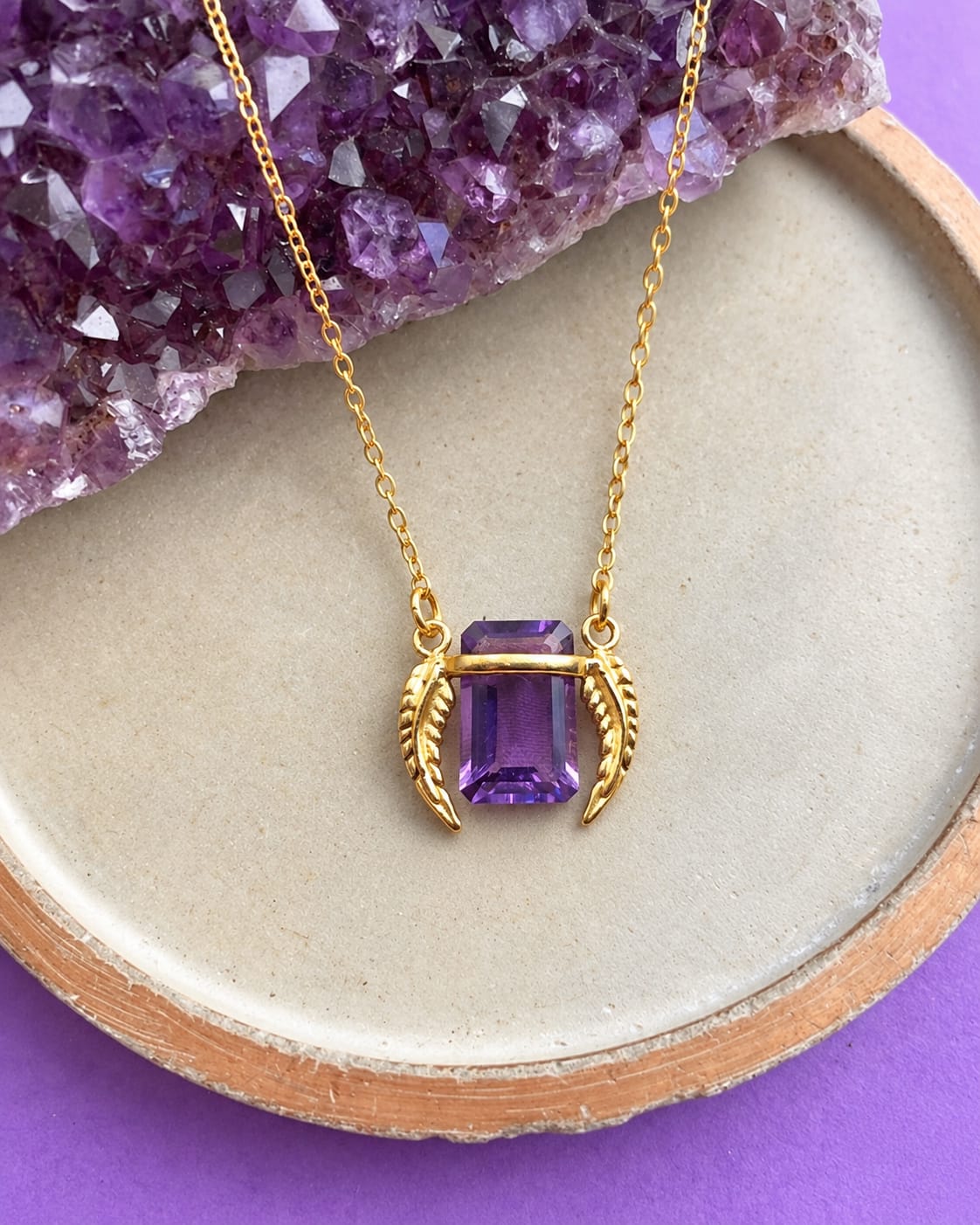 Purple rectangular pendant with gold-coloured leaf details, resting on a ceramic tray beside a purple crystal cluster.